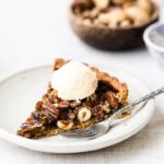 A slice of caramel nut tart topped off with a scoop of vanilla ice cream is served on a plate.