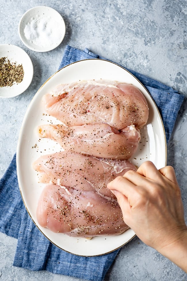 Chicken breasts are being sprinkled with salt and pepper by a woman 