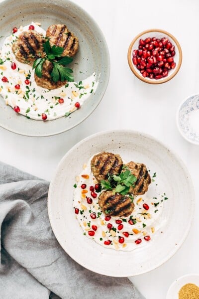 Two bowls filled with yogurt tahini sauce and Turkish meatballs garnished with pomegranate seeds and pine nuts.