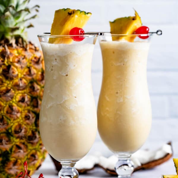 Two glasses of virgin pina colada garnished with maraschino cherries and pineapple wedges.